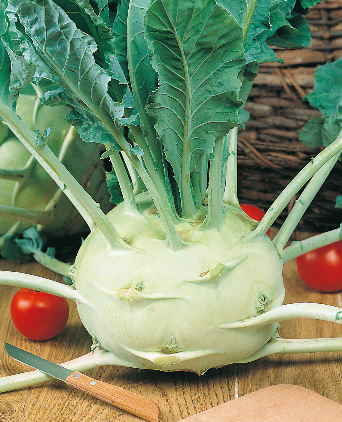 Nahaufnahme eines frischen grünen Kohlrabis mit Blattgrün auf einem Holzbrett, daneben zwei Tomaten und ein kleines Küchenmesser