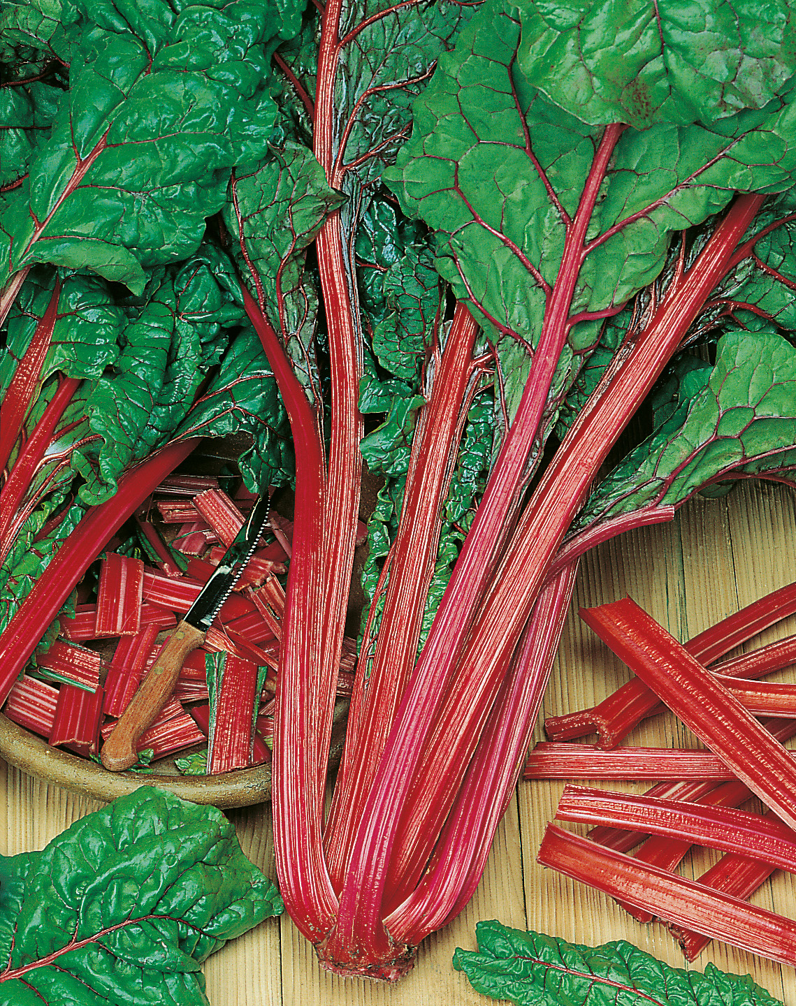 91490_Mangold Rhubarb Chard Nahaufnahme eines Bunds roter Mangold mit glänzenden roten Stielen, grünen Blattadern, geschnittenen Stücken und Messer auf Holzuntergrund