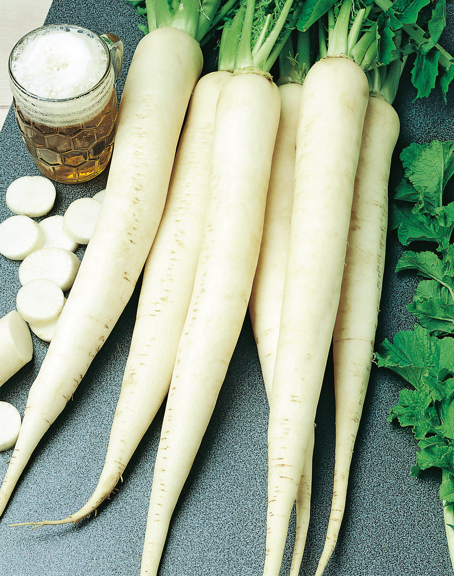 92650_Rettich Sommerwunder Frische Daikon-Rettiche (weißer Rettich) mit grünem Blattwerk und geschnittenen Scheiben, liegend auf dunklem Untergrund neben einem Bierglas