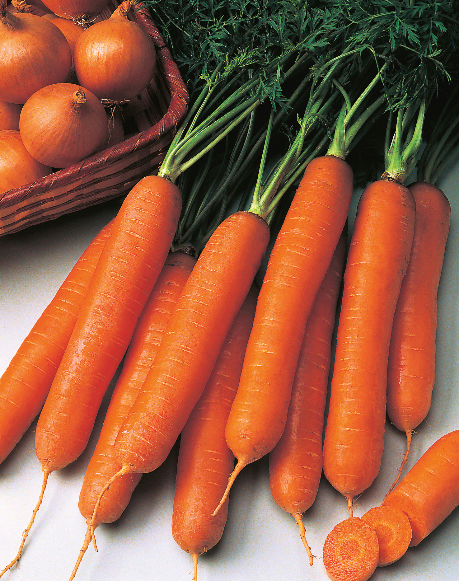 Nahaufnahme mehrerer frischer orangefarbener Karotten mit grünen Blättern, daneben Zwiebeln in einem geflochtenen Korb