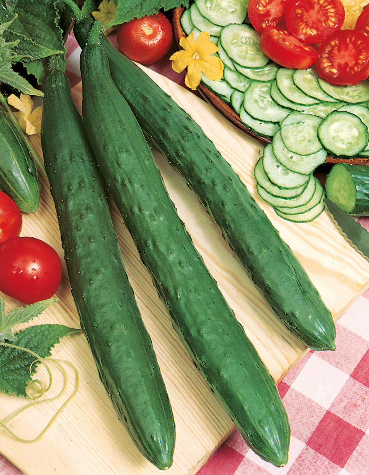 90690_Freilandgurken Tasty Green F1 Drei lange Schlangengurken auf Holzbrett, daneben geschnittene Gurkenscheiben, Tomaten und gelbe Gurkenblüten