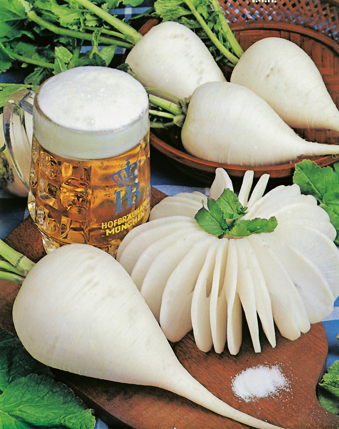 92670_Rettich Ovale blanc de Munich Weißer Daikon-Rettich: mehrere ganze Knollen und eine fein aufgeschnittene Knolle auf einem Holzbrett mit frischen Blättern, einer Prise Salz und einem Bierkrug im Hintergrund