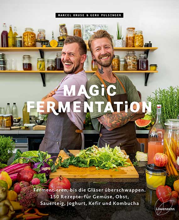 Zwei lächelnde Männer mit Schürzen stehen Rücken an Rücken in einer Küche; auf dem Tisch vorn ein Schneidebrett mit Salat, Obst und Gemüse; im Hintergrund Regale mit Einmachgläsern und fermentierten Lebensmitteln.