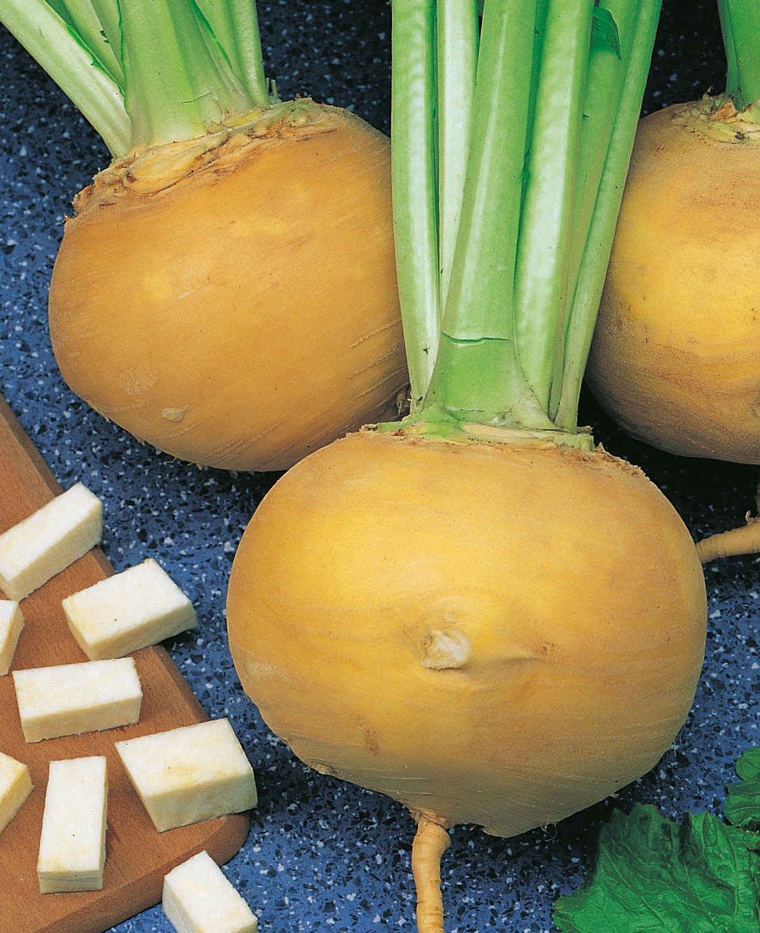 Nahaufnahme von ganzen gelben Steckrüben (Rutabaga) mit grünen Stielen; daneben gewürfelte Stücke auf einem Holzbrett auf blauem Tisch