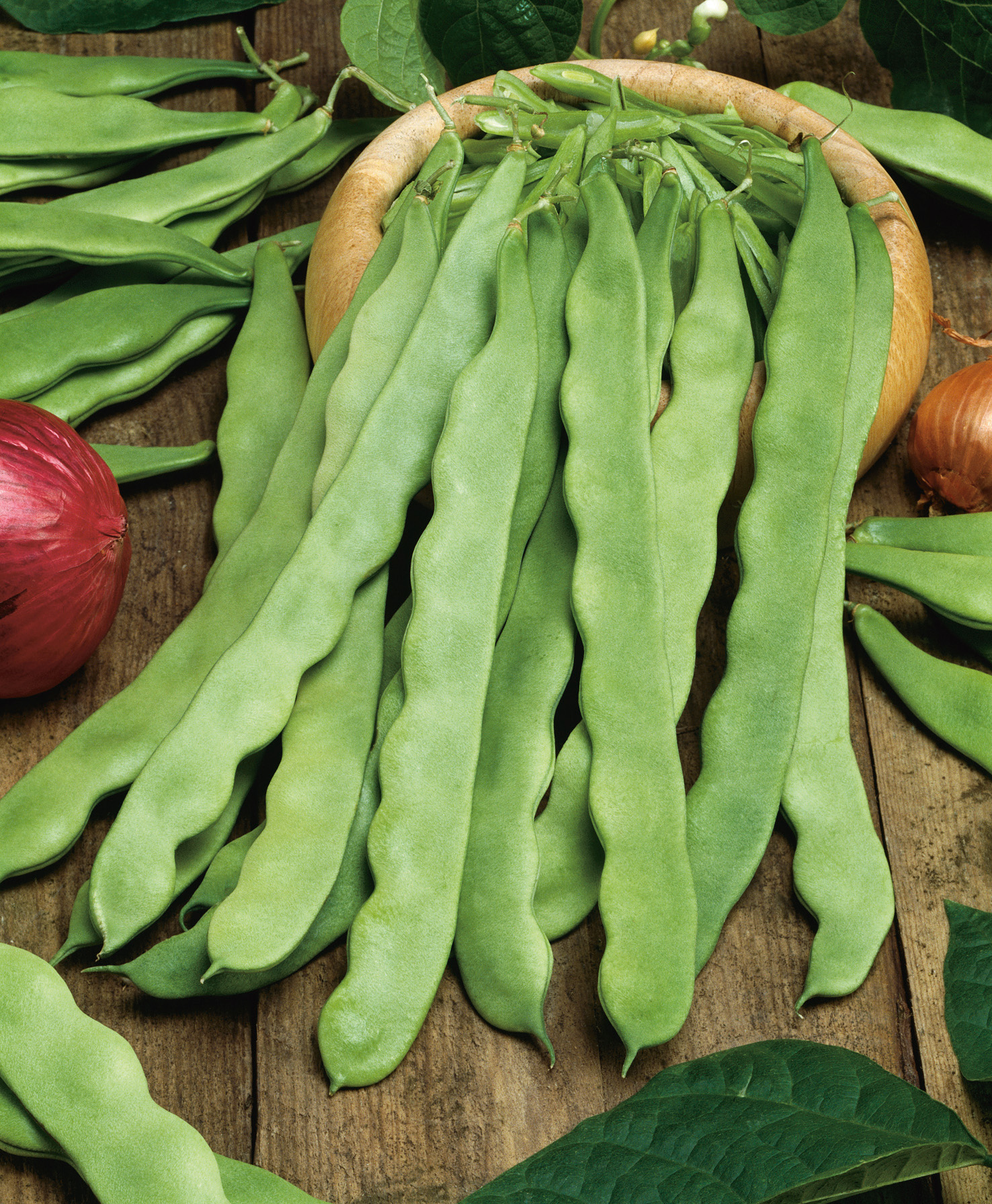 Frische, lange flache grüne Gartenbohnen in einer Holzschale auf rustikalem Holztisch, nebeneinander gelegen, mit roten und gelben Zwiebeln im Hintergrund