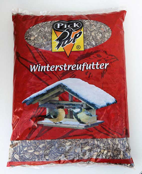 Packung Pick Pip Winterstreufutter in roter Plastikverpackung mit sichtbarem Fenster, gefüllt mit Sonnenblumenkernen; aufgedrucktes Motiv eines verschneiten Futterhauses mit zwei Meisen