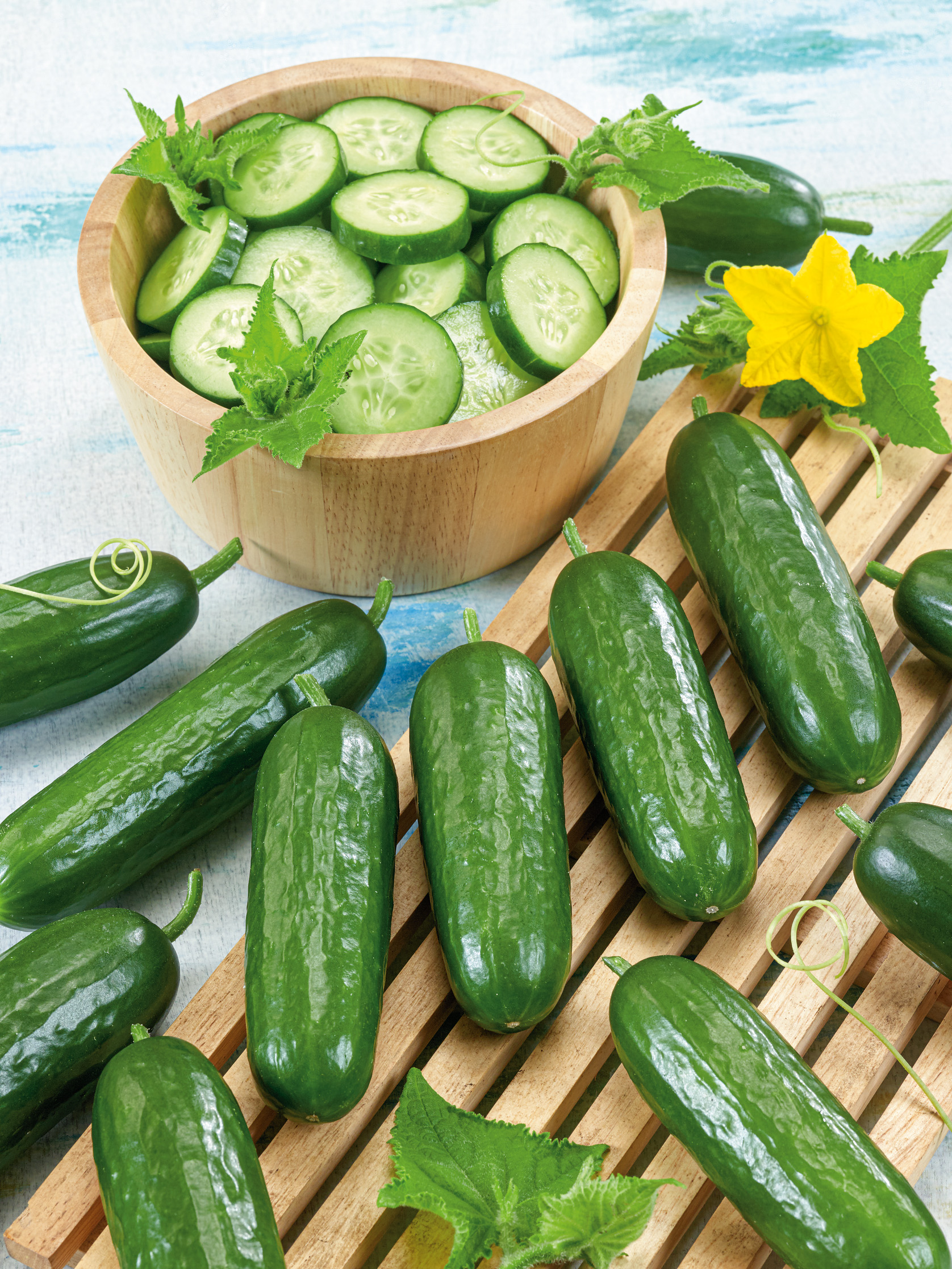 Mehrere glänzende grüne Snackgurken auf einer Holzlatte, daneben eine Holzschüssel mit frisch geschnittenen Gurkenscheiben, eine gelbe Gurkenblüte und grüne Blätter