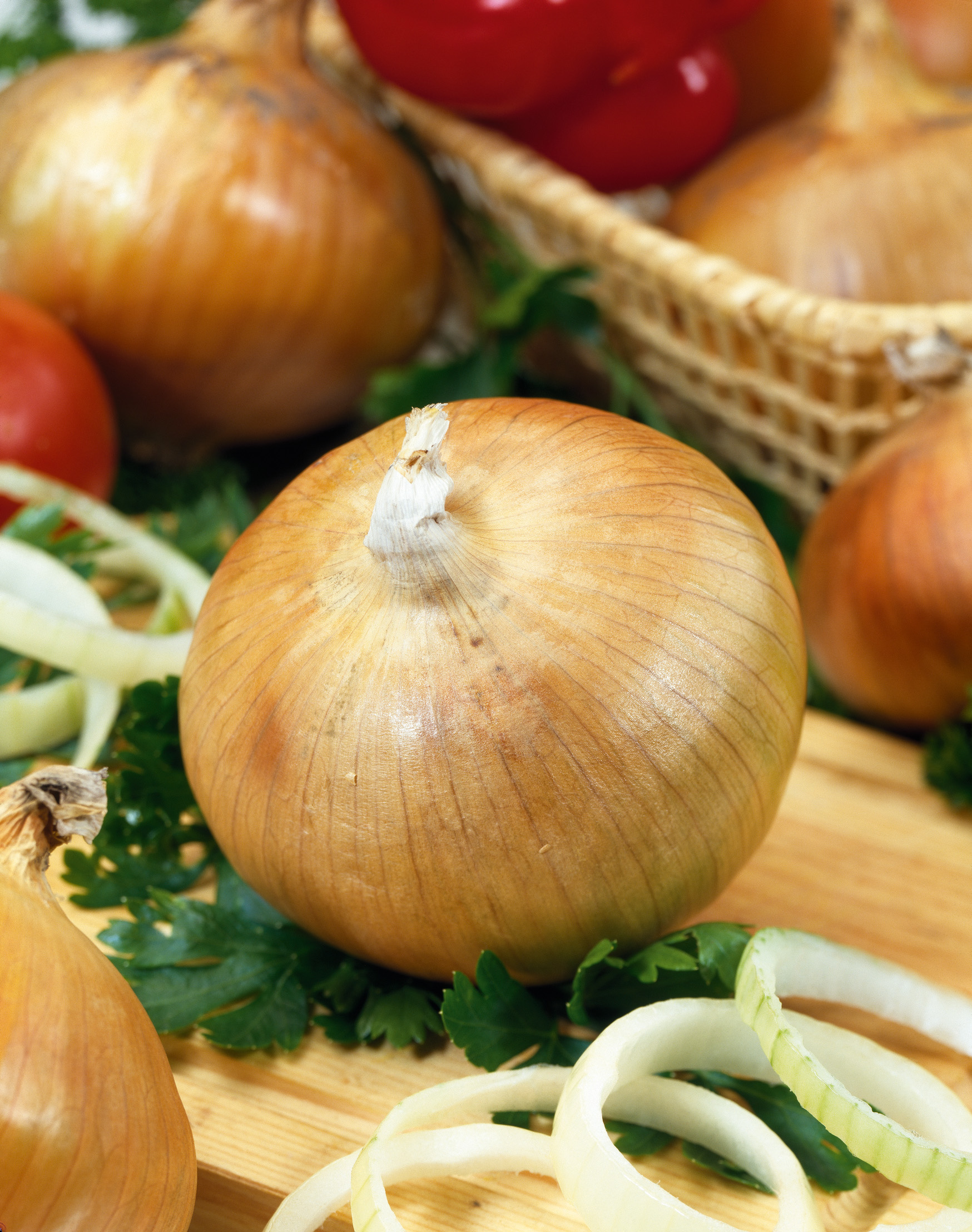 Nahaufnahme einer gelben Zwiebel auf einem Holzbrett, umgeben von Zwiebelscheiben, Petersilie und Tomaten im Hintergrund