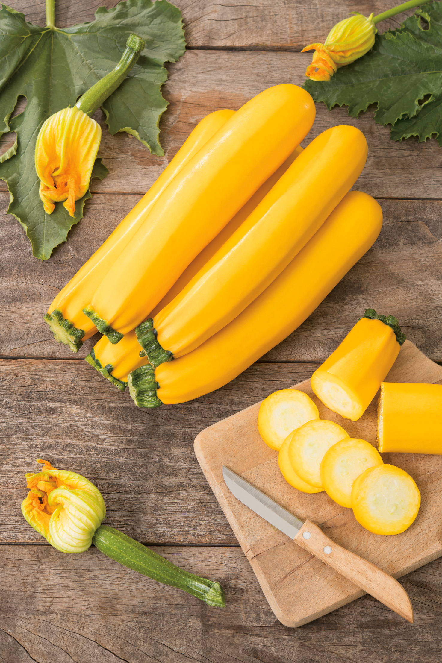 Mehrere helle gelbe Zucchini auf rustikalem Holzuntergrund, einige geschnitten auf einem Holzbrett neben Messer, mit Zucchiniblüten und Blättern – Nahaufnahme
