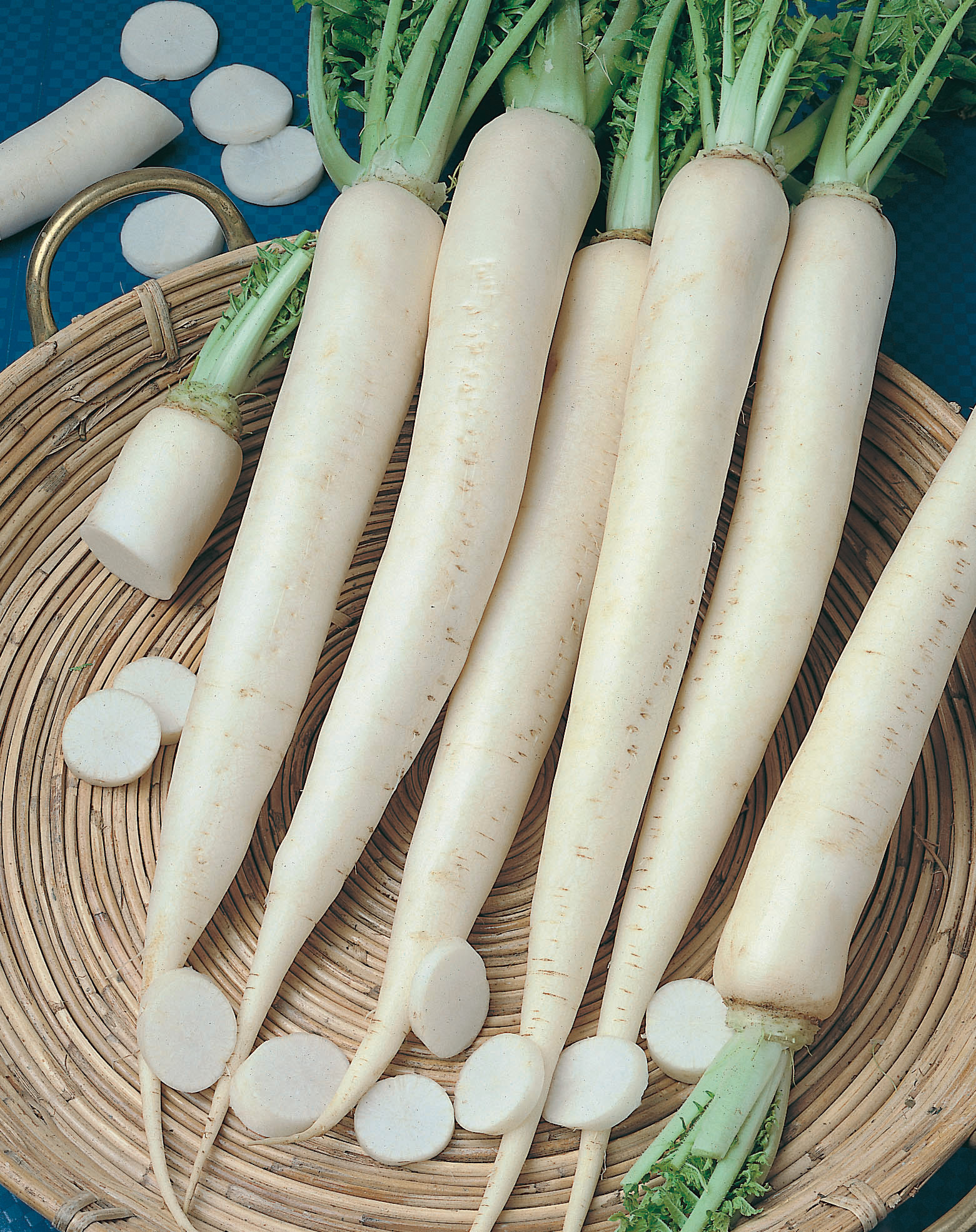 92640_Rettich Neptun F1 Mehrere lange weiße Rettiche (Daikon) liegen in einem flachen Weidenkorb, einige in dünne Scheiben geschnitten, grüne Blattstiele sichtbar.