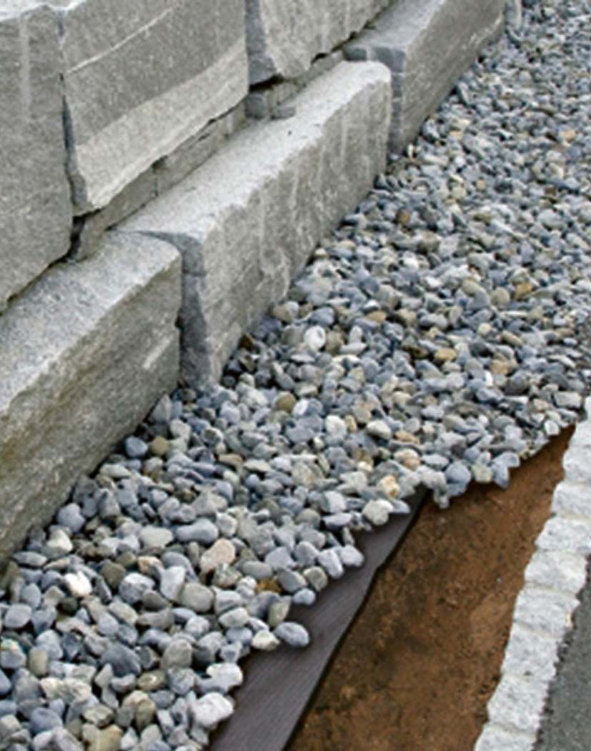 Nahaufnahme grauer Zierkiesel (Splitt) auf Unkrautvlies neben gestapelter Natursteinmauer und gepflastertem Rand