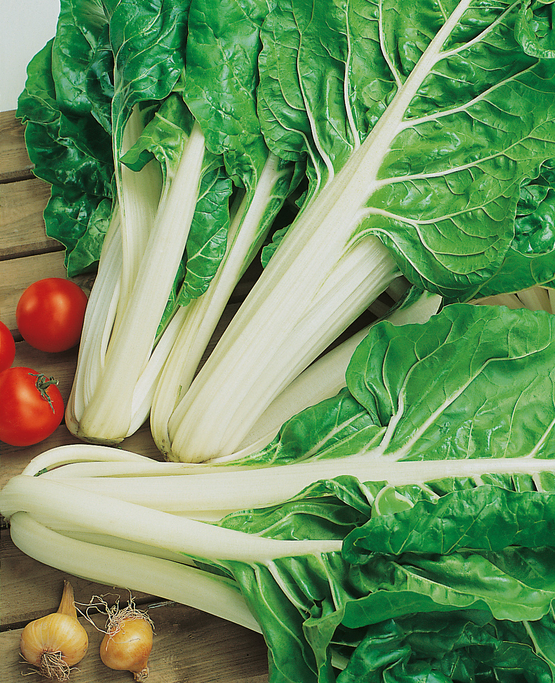 Nahaufnahme von frischem weißstieligem Mangold mit leuchtend grünen Blättern auf Holzuntergrund, daneben Kirschtomaten und Schalotten.