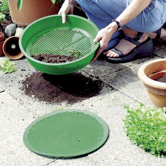 Person siebt Erde mit rundem grünem Plastiksieb auf Terrasse neben Blumentöpfen