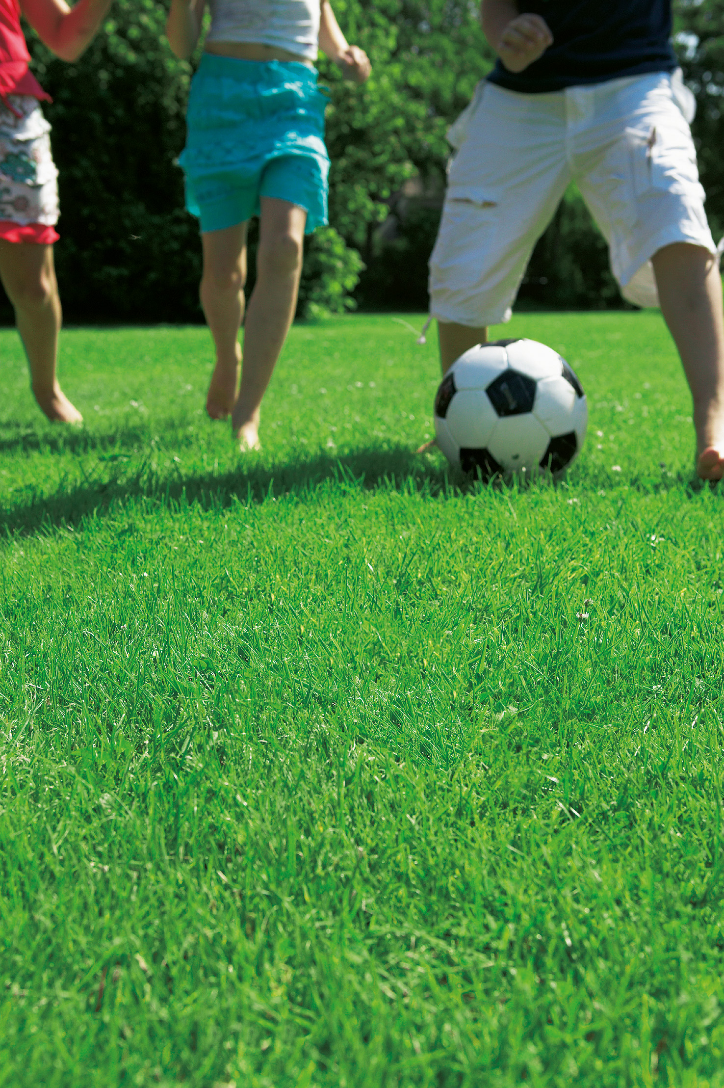 Drei Kinder laufen barfuß über grünen Rasen und kicken einen schwarz-weißen Fußball